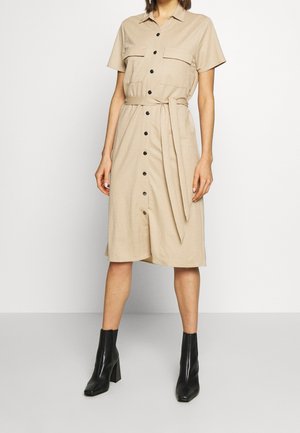 Woman wearing a beige button-up shirt dress with short sleeves, two chest pockets, a tied waist belt, and black ankle boots.