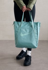 Persoon in zwarte legging en groene jas houdt een turquoise Nike-tas vast, draagt zwarte Nike-sneakers en witte Nike-sokken.