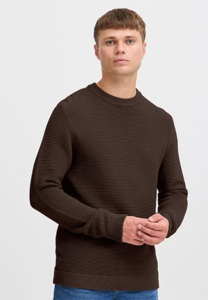 Jeune homme aux cheveux bruns courts portant un pull à manches longues marron foncé texturé et un jean bleu sur un fond clair uni.