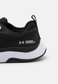Scarpa sportiva nera con parte superiore in mesh, pannelli laterali testurizzati, suola bianca e logo Under Armour sul tallone. Presenta una linguetta di trazione.