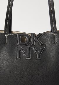 Black leather tote bag featuring a textured surface, white stitching, and bold DKNY logo in black. Open top with dual handles.