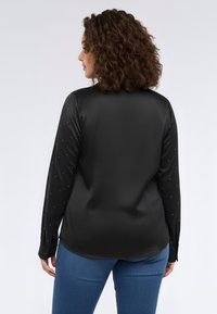 Blusa nera a maniche lunghe con una texture liscia, dettagli di scintillio sottili e orlo arrotondato. Abbinata a jeans blu.