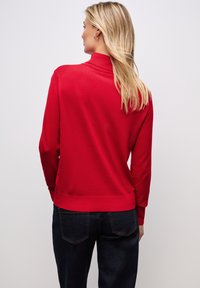 Pullover rosso con colletto alto e maniche lunghe, caratterizzato da una superficie liscia e orli a costine. Indossato con jeans scuri.