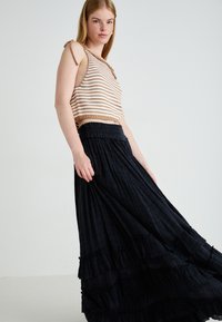 Jeune femme portant un crop top en tricot rayé sans manches et une longue jupe noire fluide, se tenant devant un fond clair uni.