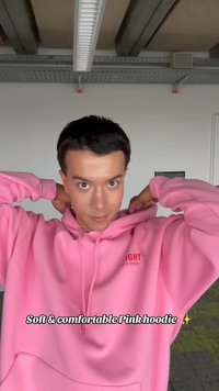 Ung mann som justerer en knallrosa hettegenser innendørs, med taklampe og industrielt tak i bakgrunnen.