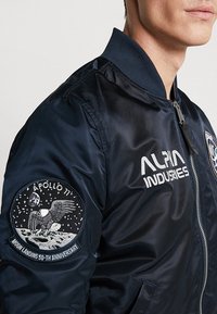 Chaqueta bomber azul marino con cierre de cremallera, cuello acanalado y un parche del aniversario del Apolo 11 en la manga izquierda. Tejido liso y brillante.