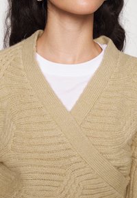 Femme portant un cardigan beige en tricot sur un t-shirt blanc à encolure ronde, avec de longs cheveux bruns et des boucles d'oreilles anneaux.