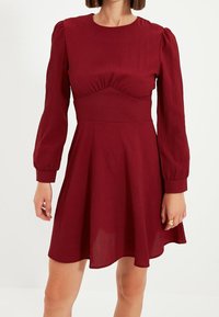 Burgundy klänning med figursydd överdel, puffärmar och utsvängd kjol. Tillverkad av strukturerad tyg med en slät finish.