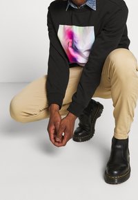 Sudadera negra con un gráfico abstracto colorido y un logo en el frente, combinada con pantalones beige y botas negras con costuras amarillas.