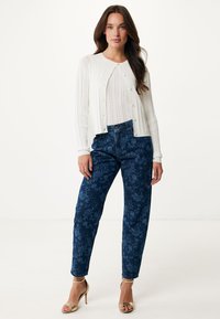 Biała prążkowana kardiganka z zapięciem na guziki, w połączeniu z niebieskimi dżinsami w kwiatowy wzór i złotymi sandałami na wysokim obcasie z paseczkami.