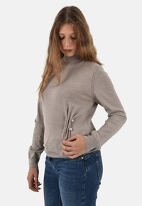 Maglioncino beige chiaro con collo alto, cuciture testurizzate e tre bottoni decorativi sul lato. Abbinato a jeans blu.