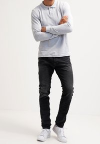 Person stands with arms crossed wearing a light gray long-sleeve polo, black jeans, and white sneakers against a plain white background.