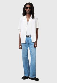 Camicia bianca a maniche corte con motivo testurizzato, abbinata a jeans in denim blu chiaro a gamba larga e scarpe slip-on nere. Indossati occhiali da sole e collana.