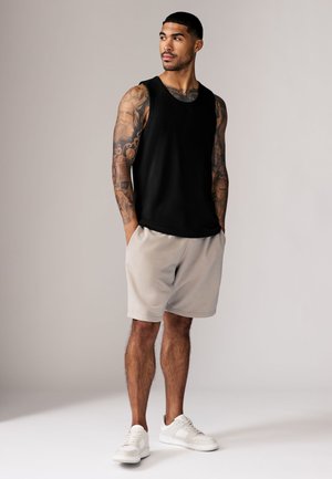 Muž s potetovanými rukami, oblečený v čiernom tielku, béžových šortkách a bielych teniskách, stojí s rukami v vreckách a pozerá sa bokom.