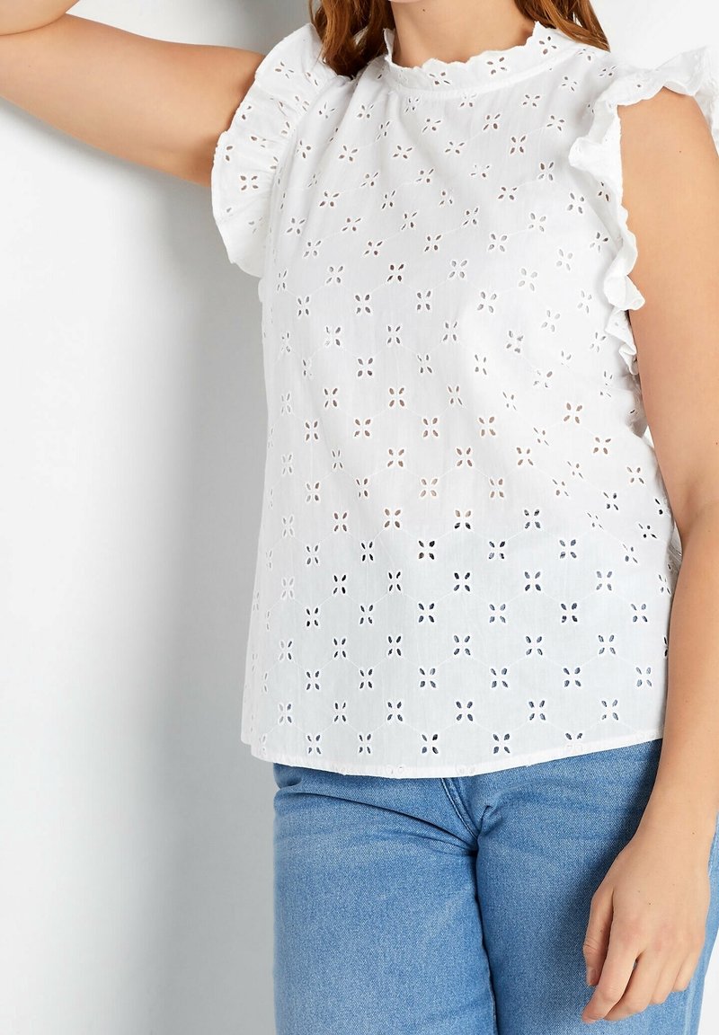 Femme portant un haut blanc sans manches en dentelle ajourée avec des épaules à volants et un jean bleu clair, bras appuyé contre un mur.