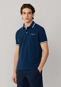 Giovane uomo con polo blu navy con colletto e maniche a righe bianche, posando con una mano in tasca davanti a uno sfondo semplice.