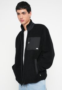 Juodas vilnos švarkas su aukštu apykakliu, turintis krūtinės kišenę su užtrauktuku, kontrastingomis juodo audinio detalėmis ir laisvo silueto.