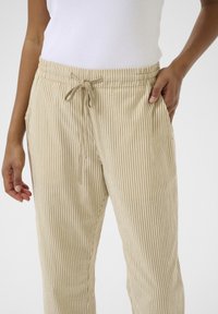 Pantalon en velours côtelé beige à rayures verticales Beanie, doté d'une taille élastique et d'un cordon de serrage, présenté avec les mains dans les poches.