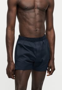 Αθλητικός σκουρόχρωμος άνδρας που φοράει σκούρο μπλε σορτς με ελαστική μέση, στέκεται με τα χέρια χαλαρά σε απλό φωτεινό φόντο.