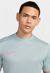Jasnoniebieska koszulka sportowa z gładkiego materiału, z okrągłym dekoltem. Posiada różowe logo Nike na piersiach. Krótkie rękawy, dopasowany krój.