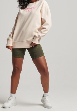 Frau trägt einen beigen übergroßen Pullover mit pinkem Logo, olivgrüne Radlerhosen, weiße Socken und weiße Turnschuhe, steht mit gespreizten Beinen.