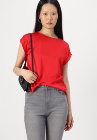 Top rojo de manga corta con un corte relajado, combinado con jeans ajustados grises. Un bolso cruzado negro añade contraste. Se puede apreciar la textura suave de la tela.