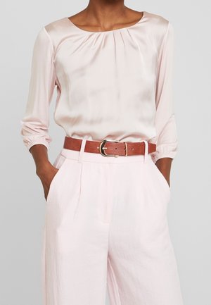 Persona que lleva una blusa de satén rosa pálido con escote fruncido, pantalones de cintura alta a juego y un cinturón de cuero marrón con hebilla dorada.