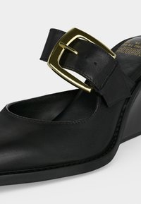 Γυναικείο μαύρο δερμάτινο παπούτσι με μυτερή μύτη, χοντρό τακούνι και μεγάλο χρυσό αγκράφα στο πάνω μέρος του ποδιού.