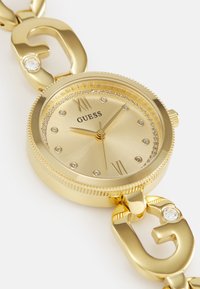 Orologio dorato con cassa rotonda, numeri romani e dettagli in cristallo attorno al quadrante. Bracciale a maglie con dettagli hardware firmati.