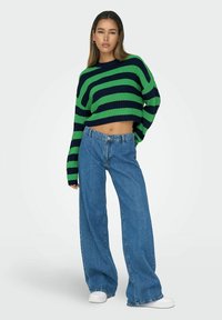 Adīkts ar adītu virsu ar drosmīgiem zaļiem un tumši zilajiem svītrām, apvienots ar augsta vidukļa platām denim biksēm un baltām sporta apavicēm.
