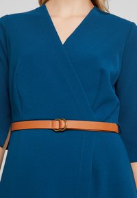 Klänning i solid teal-tyg med korta ärmar, utrustad med ett bälte i beige konstläder med detaljer i guldton.