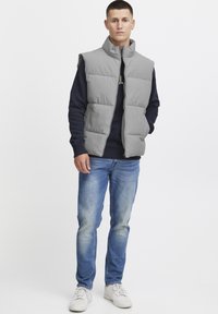 Graue Steppweste mit hohem Kragen, Reißverschluss und gesteppter Optik. Getragen über einem marineblauen Pullover, mit blauen Jeans und weißen Sneakers.