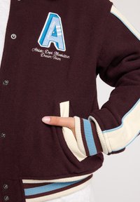 Maroon varsity jacket with a blue embroidered 'A' logo, cream accents, striped cuffs, and snap button closure. Soft, textured fabric.