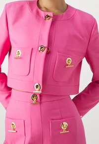 Giacca corta rosa con bottoni rotondi in oro e due tasche frontali con simili accenti dorati. Tessuto liscio e su misura.