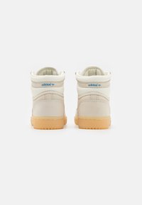 High-top Adidas sneakers in cream leather with a textured finish, featuring blue logo accents and a beige rubber sole.