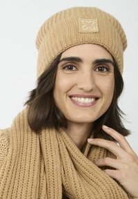 Gehaakte beanie in zacht beige met een geribbelde textuur en een vierkante logopatch, gecombineerd met een bijpassende dikke sjaal in een vergelijkbare kleur.