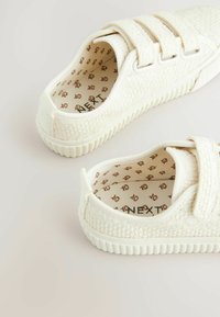 Baskets blanches avec une tige texturée, deux sangles Velcro et une semelle en caoutchouc striée. L'intérieur présente un motif floral brun.