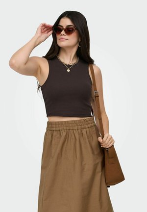 Woman adjusting cat-eye sunglasses, wearing sleeveless black crop top, brown high-waist skirt, gold necklace, hoop earrings, and carrying brown shoulder bag.