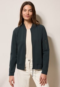 Giacca bomber verde scuro in morbido tessuto con tasca laterale con zip, polsini a coste e chiusura frontale con zip. Indossata sopra un top chiaro.