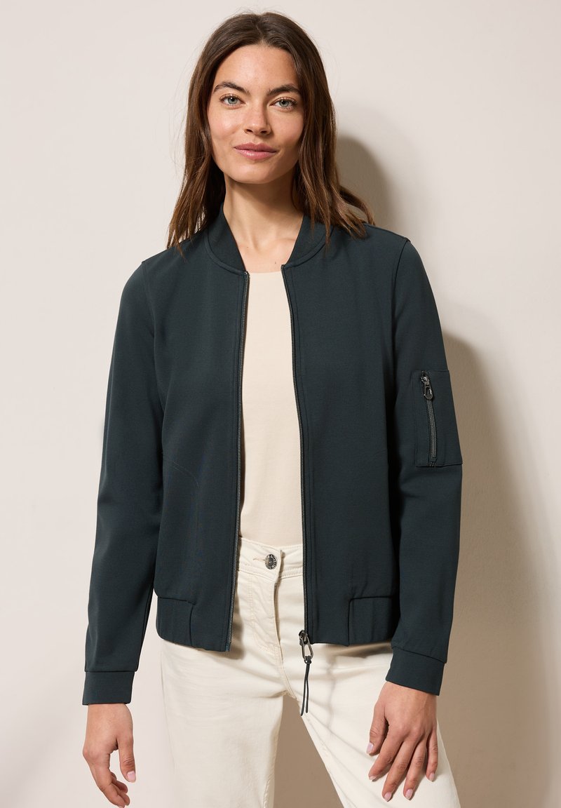 Giacca bomber verde scuro in morbido tessuto con tasca laterale con zip, polsini a coste e chiusura frontale con zip. Indossata sopra un top chiaro.