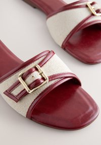 Sandales slides en cuir rouge avec des brides en tissu beige et des boucles carrées dorées, design à bout ouvert sur une surface claire.