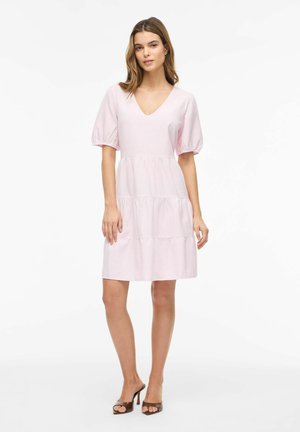 Žena v svetloružových šatách po kolená s nadýchanými rukávmi a výstrihom do V, v kombinácii s hnedými sandálmi na opätku, stojaca na bielom pozadí.