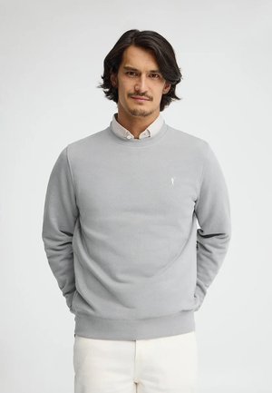 Uomo con capelli neri che indossa una felpa grigio chiaro sopra una camicia bianca, in piedi con le mani dietro la schiena davanti a uno sfondo bianco semplice.