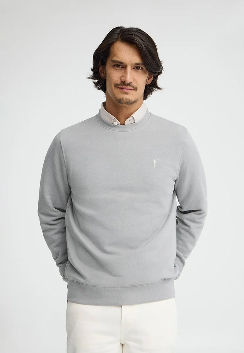 Uomo con capelli neri che indossa una felpa grigio chiaro sopra una camicia bianca, in piedi con le mani dietro la schiena davanti a uno sfondo bianco semplice.