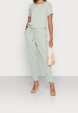 Femme portant une combinaison légère vert clair à manches courtes avec taille coulissée, tenant un sac à main beige, debout en sandales à talons hauts beige.