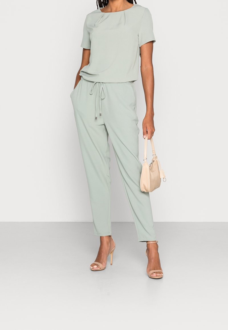 Femme portant une combinaison légère vert clair à manches courtes avec taille coulissée, tenant un sac à main beige, debout en sandales à talons hauts beige.