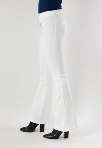 Pantaloni bianchi a zampa con una vita alta e un tessuto liscio e strutturato. Presentano una tasca posteriore singola e sono abbinati a tacchi neri.