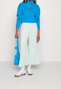 Niebieska bluza z małym logo, sparowana z jasnymi, miętowymi spodniami typu wide-leg. Trzyma niebieską torbę, nosi białe buty z brązowymi akcentami.