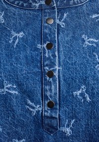 Denim košulja u plavoj boji s svijetlim apstraktnim uzorkom. Sadrži dugmad s crnim metalnim dugmadi i ovratnik.