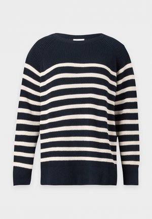 Kvinde med langt blond hår iført en sort- og hvidstribet sweater, beige vidde bukser, brune sko og holder en beige håndtaske.